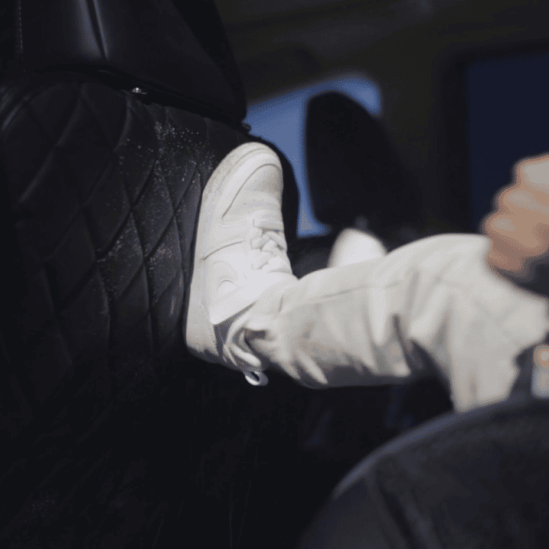 Child's white sneaker resting on a black diamond-patterned kick mat protecting the car seat back.
